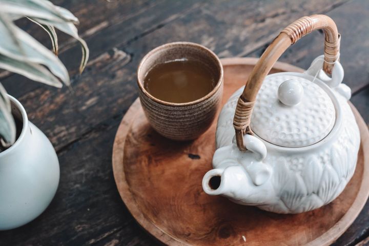 A white tea kettle and a brown cup full of tea, laying on a tray on a table beside a potted plant.