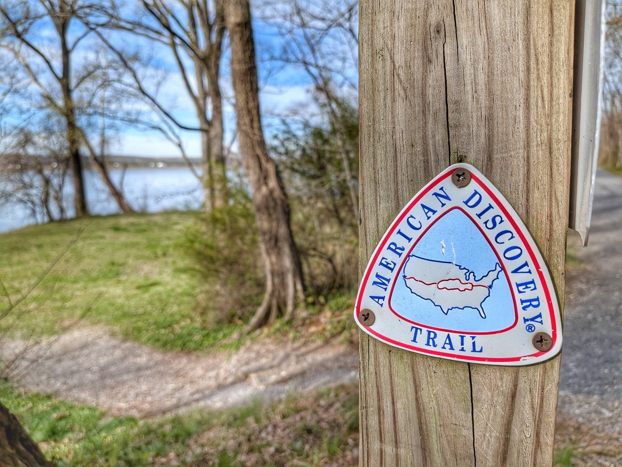 An American Discovery Trail marker on a wooden post beside the C and O Canal Towpath, with a dirt path curving left toward open water and a paved path running right