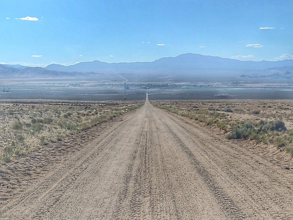 A straight dirt road running across a wide, dry sagebrush valley toward distant mountains.