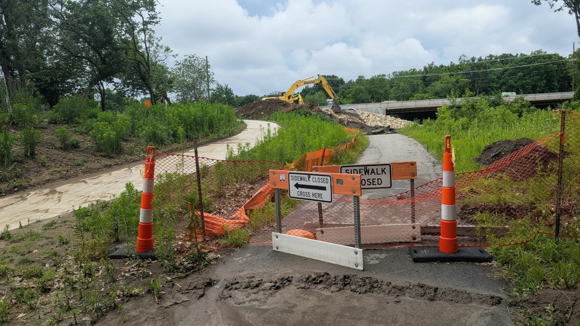 Orange barricades and a Sidewalk Closed, Cross Here sign block a paved trail beside a construction site.