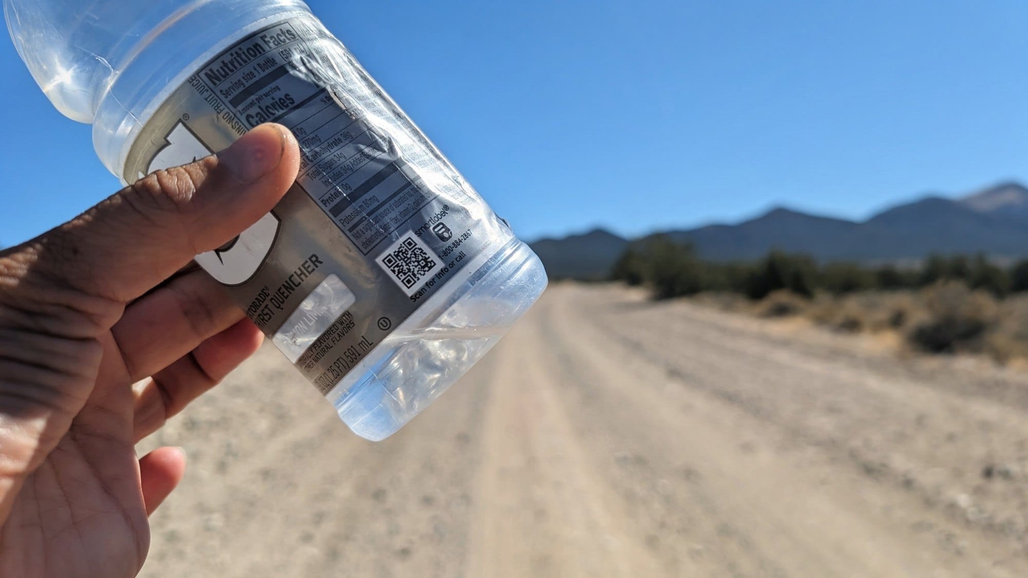 A hand holding a nearly empty plastic bottle up against a blue sky above a dirt road.