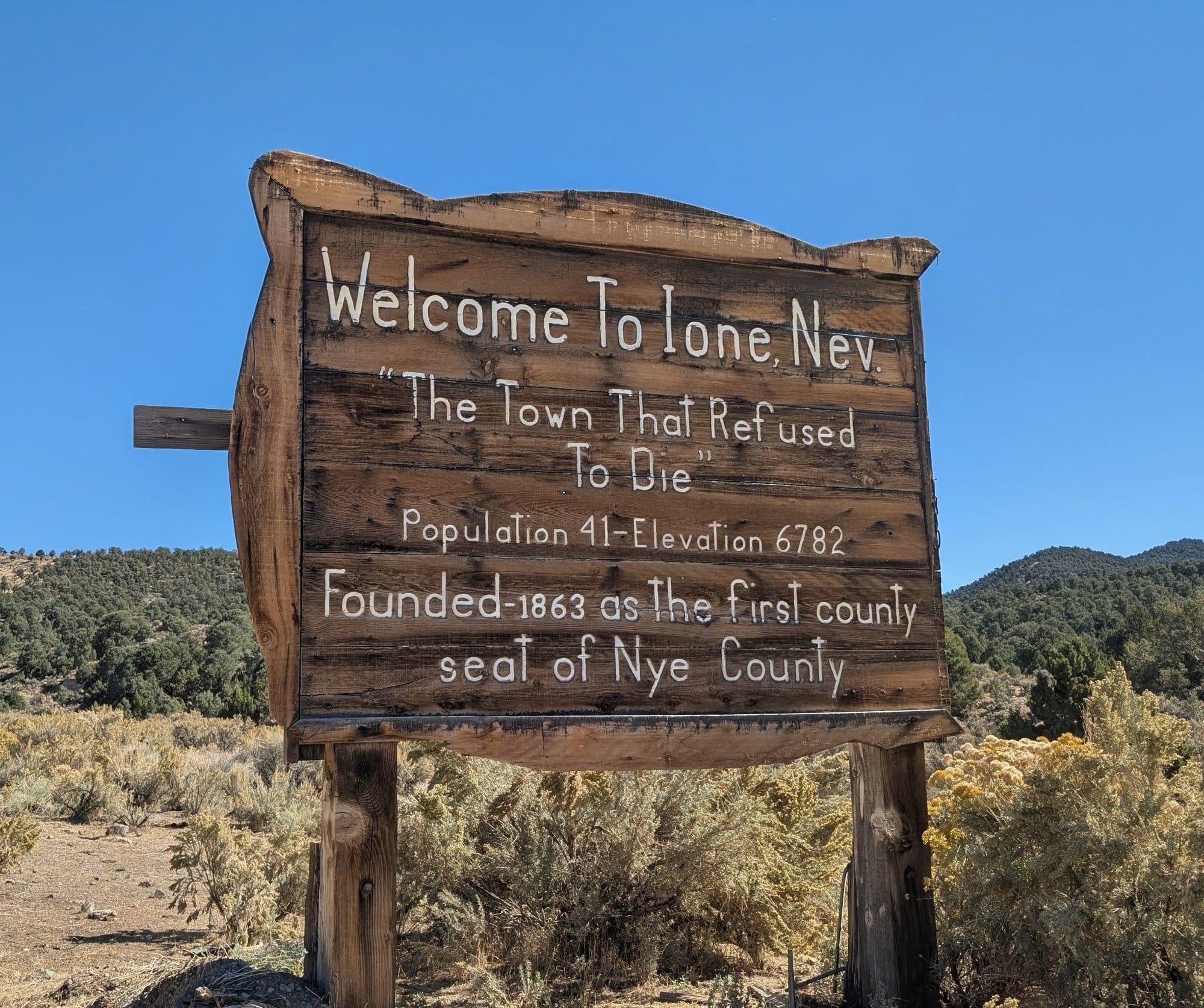 A weathered wooden sign reading Welcome to Ione, Nev, The Town That Refused to Die, Population 41, Elevation 6782, Founded 1863 as the first county seat of Nye County.
