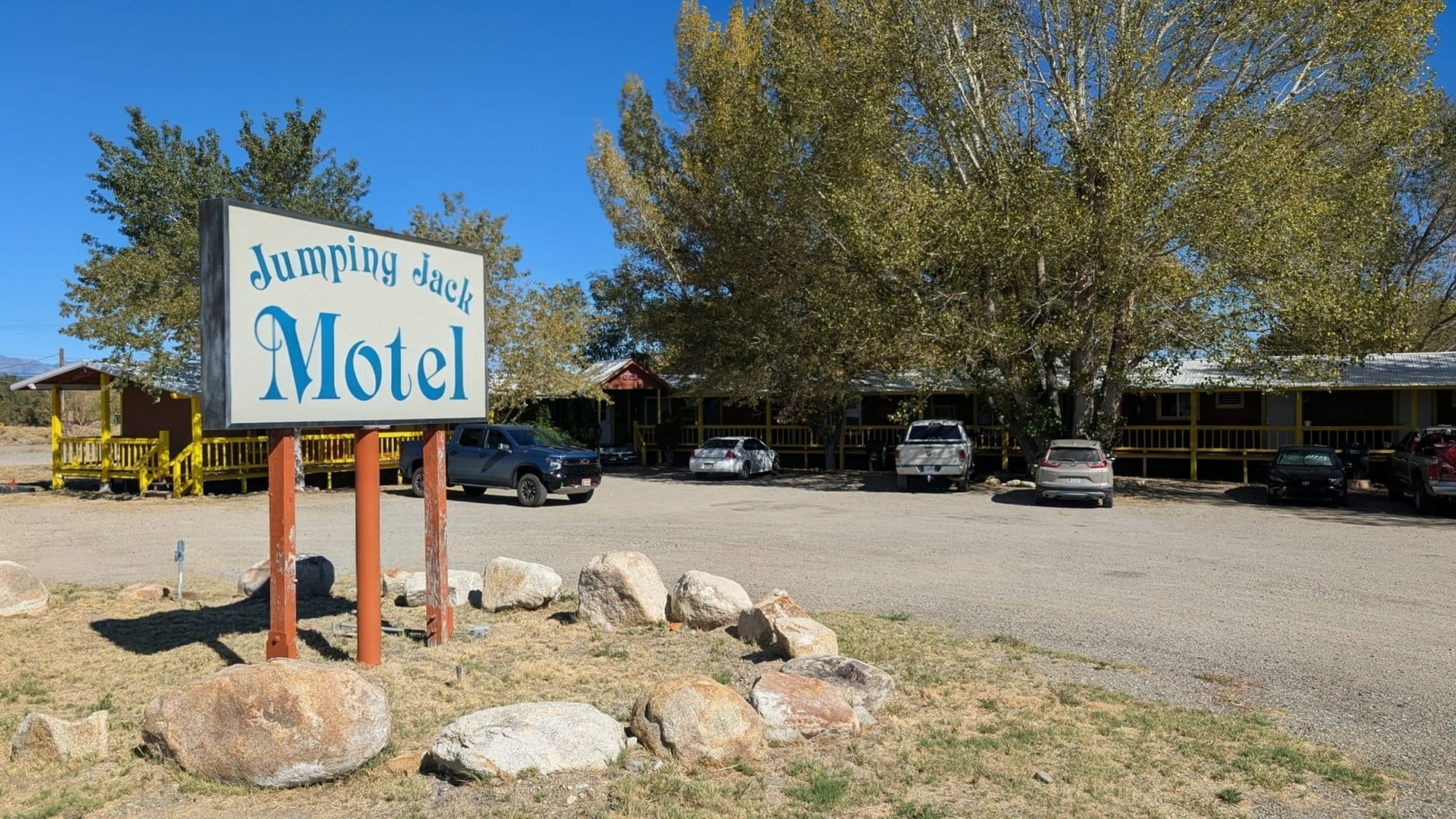 A blue and white roadside sign reading Jumping Jack Motel in front of a gravel lot, parked trucks and tall cottonwoods.