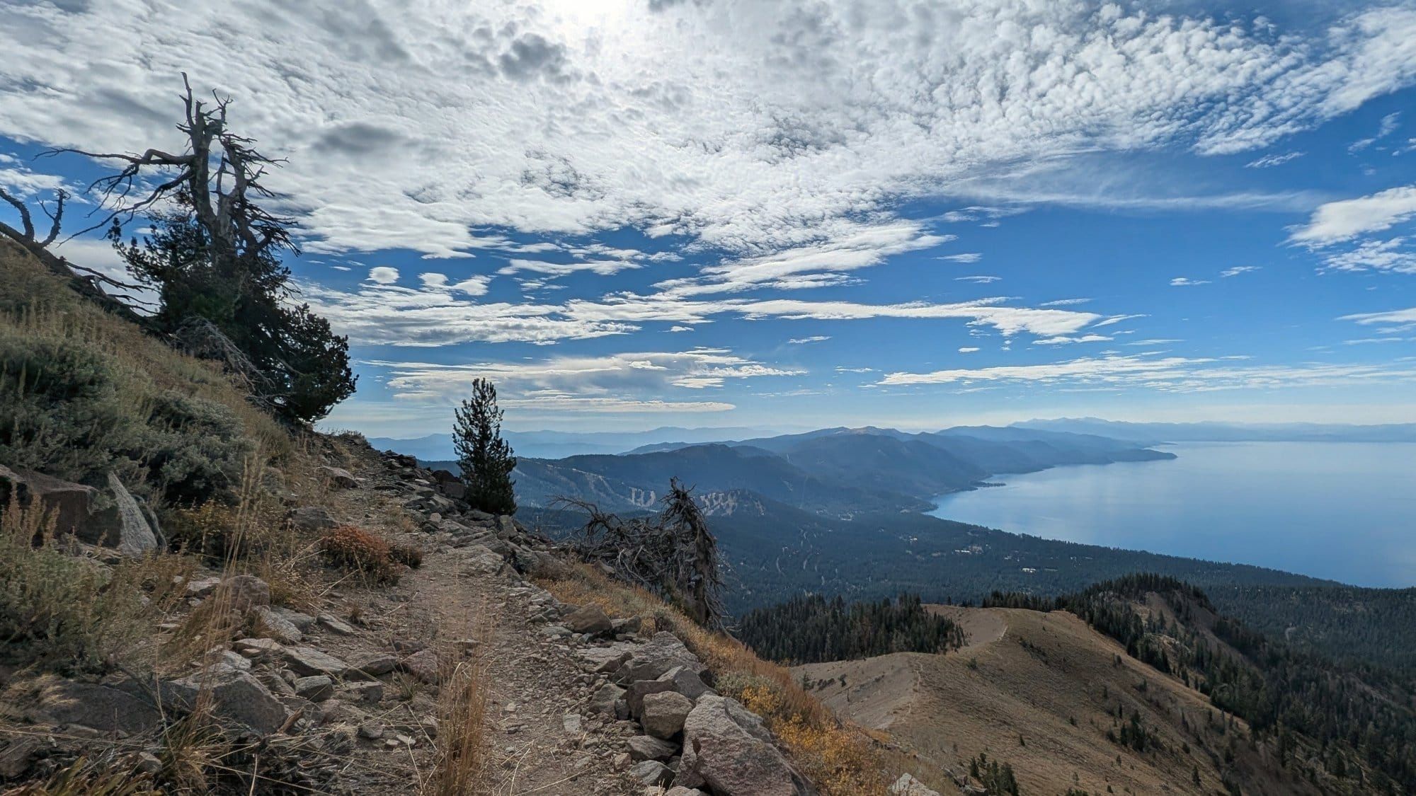 Rocky dirt trail along a dry, open ridge with wind-bent pines, Lake Tahoe's north shore far below under scattered clouds.