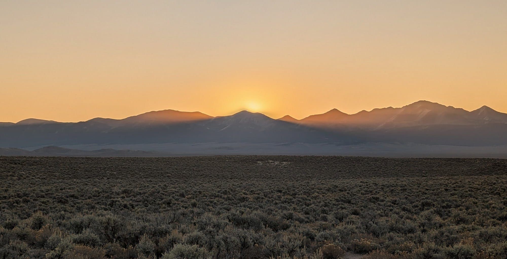 The sun rising over a jagged mountain range at the far edge of a wide, flat sagebrush valley