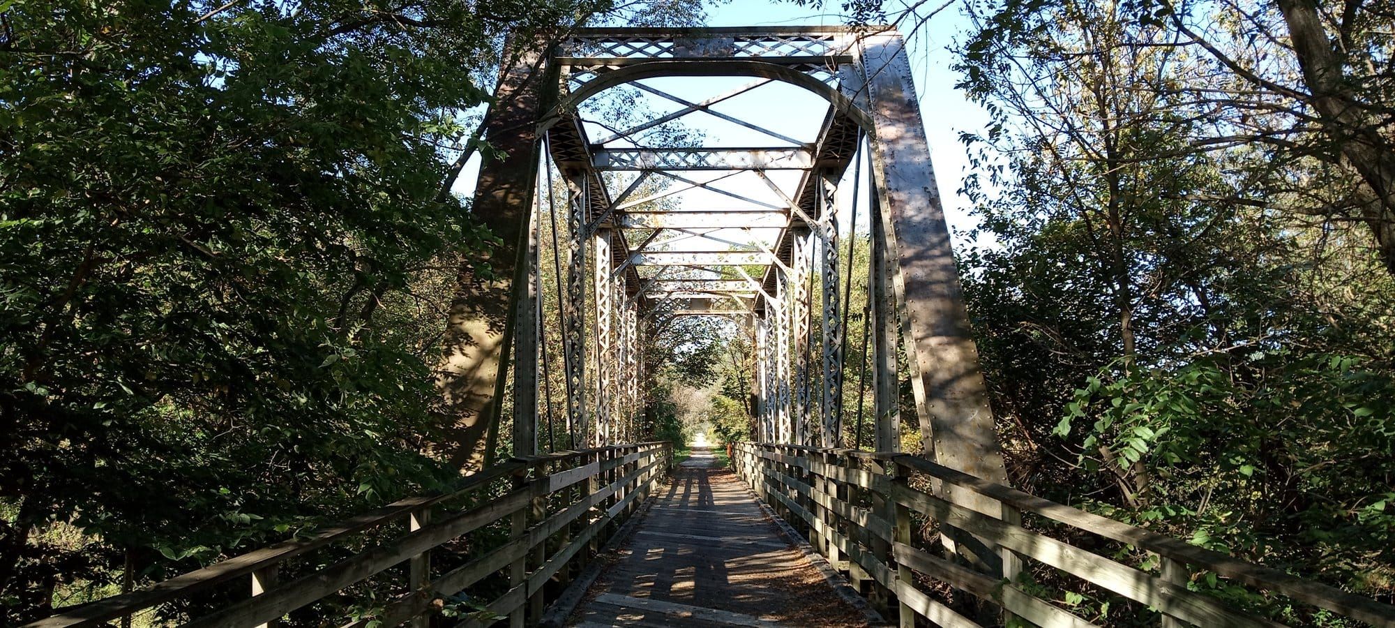 An old steel truss bridge with a wooden deck and railings, carrying the trail into dense green woods