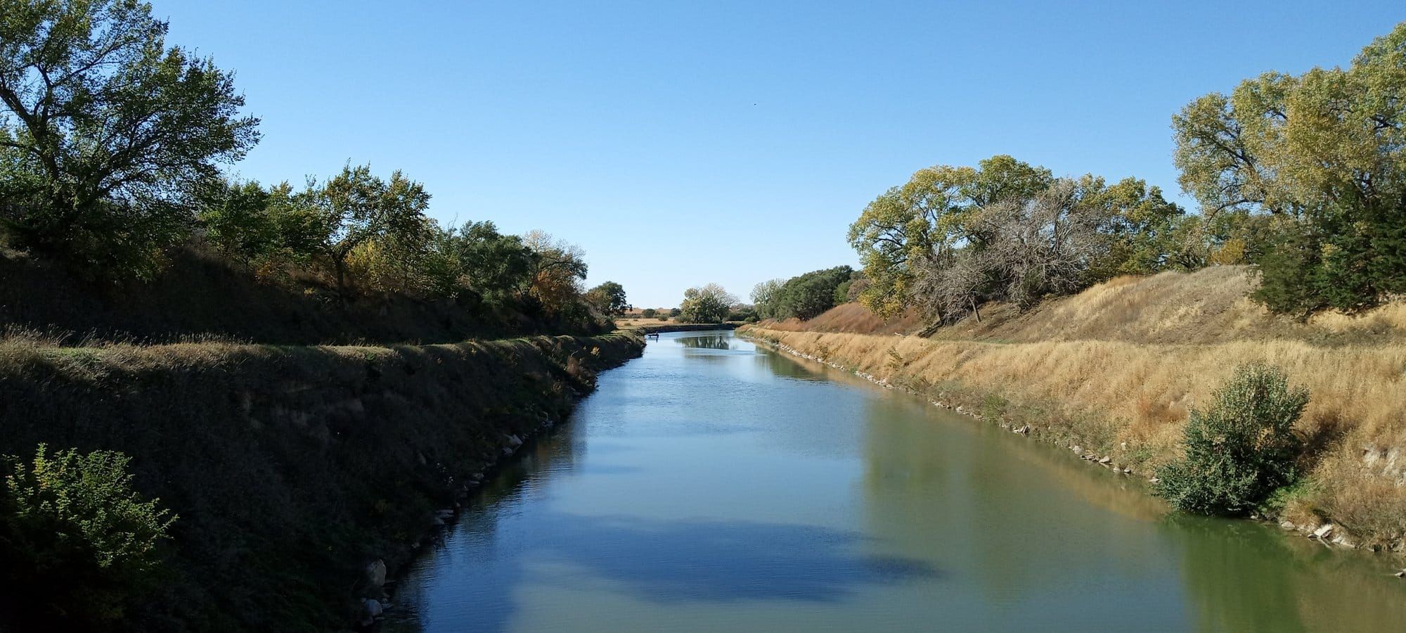 A straight irrigation canal between steep grassy banks, cottonwoods along both sides and a footbridge in the distance