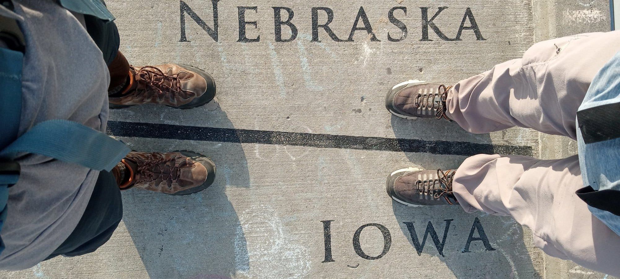 Two pairs of hiking boots straddling the inlaid state line on a concrete bridge deck, NEBRASKA on one side and IOWA on the other