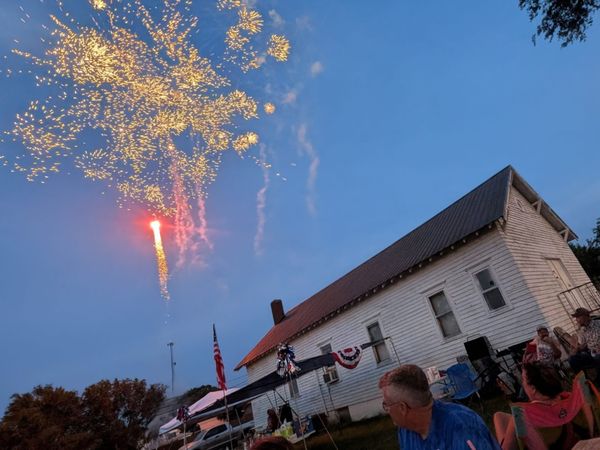 Fourth of July fireworks over a house in Ford, Kansas as people in an adjoining park look on.