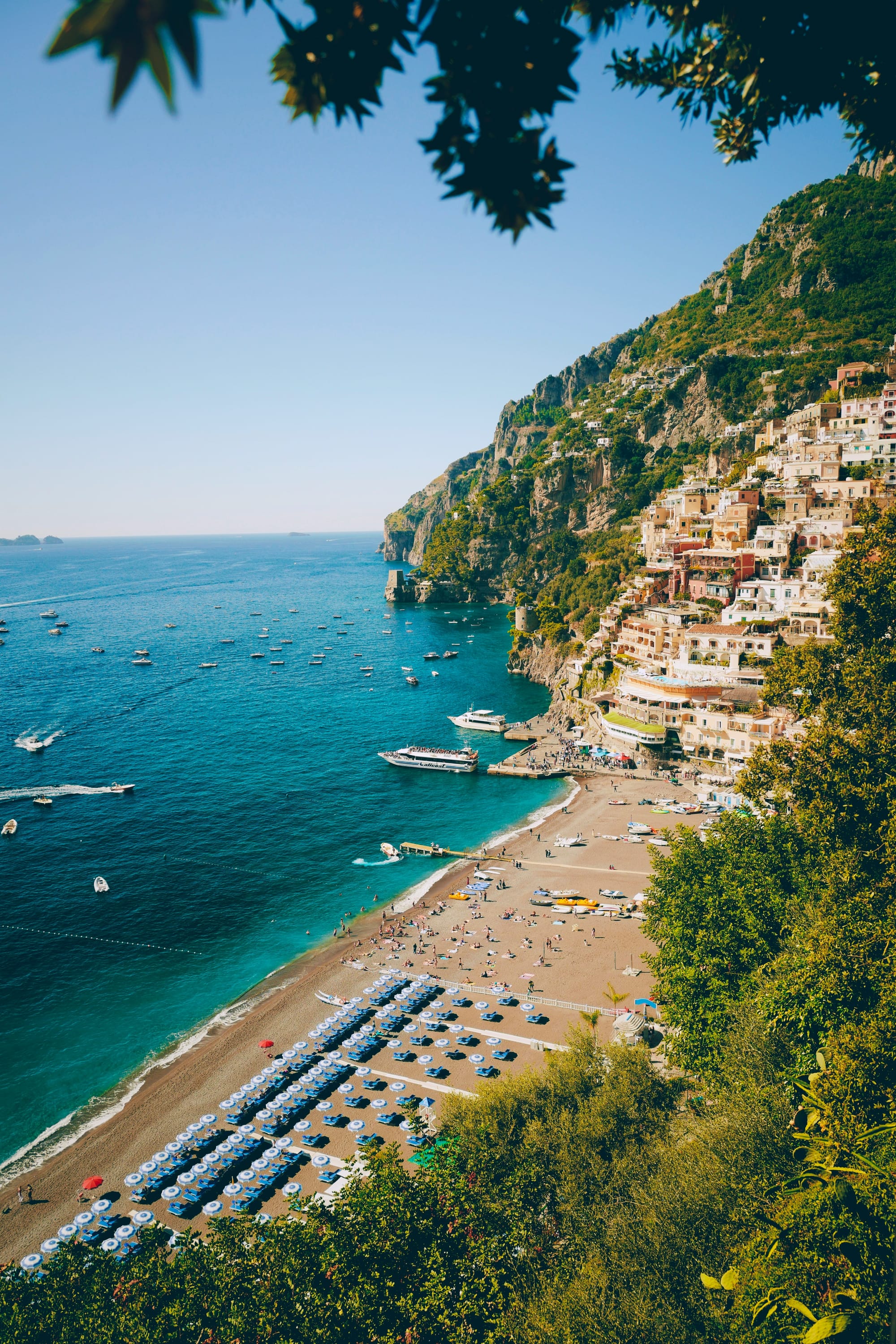 Picturesque view of the Amalfi coast with green deciduous trees in the foreground, umbrellas and sunbeds near the blue sea complete with sailboats