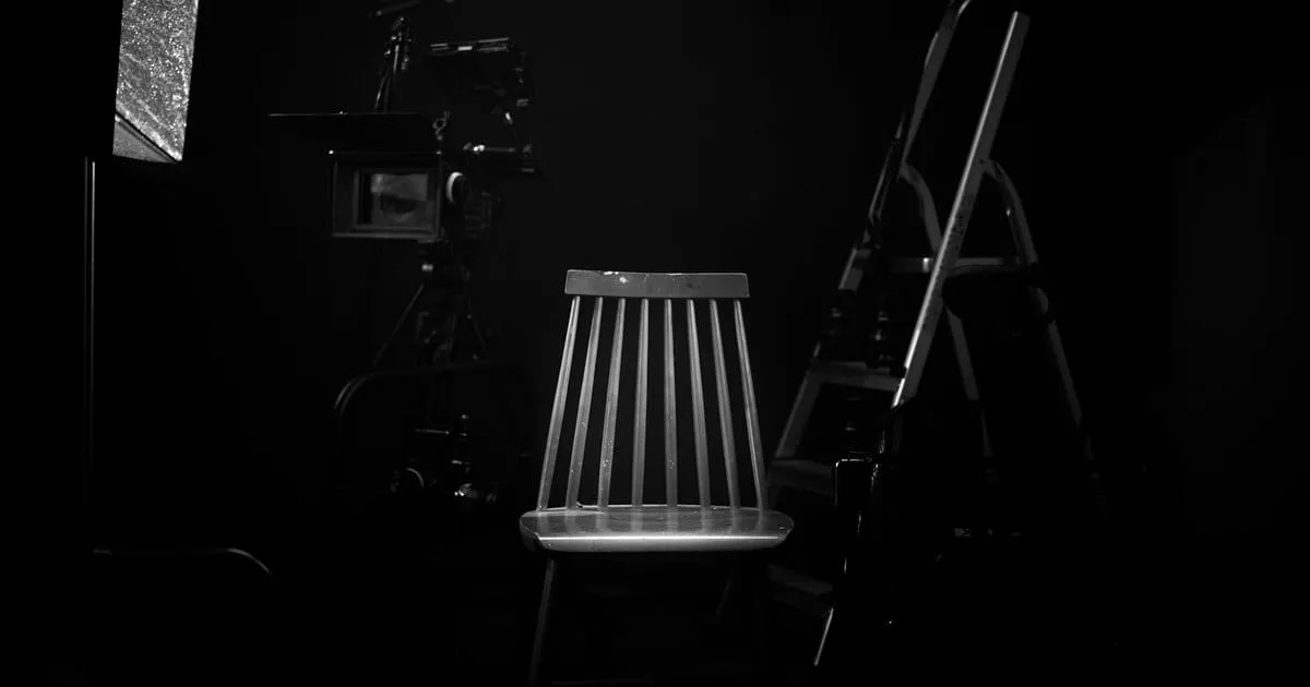 Empty wooden chair on a film studio set, surrounded by a camera, boom microphone, and lighting equipment in shadow.