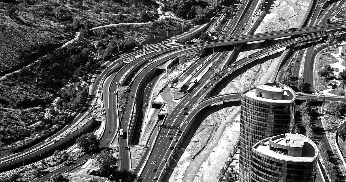 Aerial view in black and white of a multilevel highway interchange, with lanes of traffic curving past two cylindrical towers.