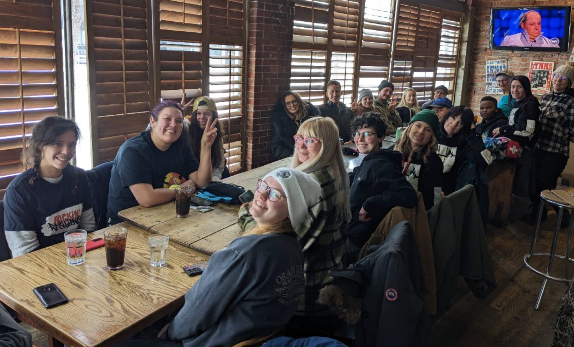 A couple dozen people sit along a table inside a cosy bar, posing for the camera. Many make peace signs. Most are smiling