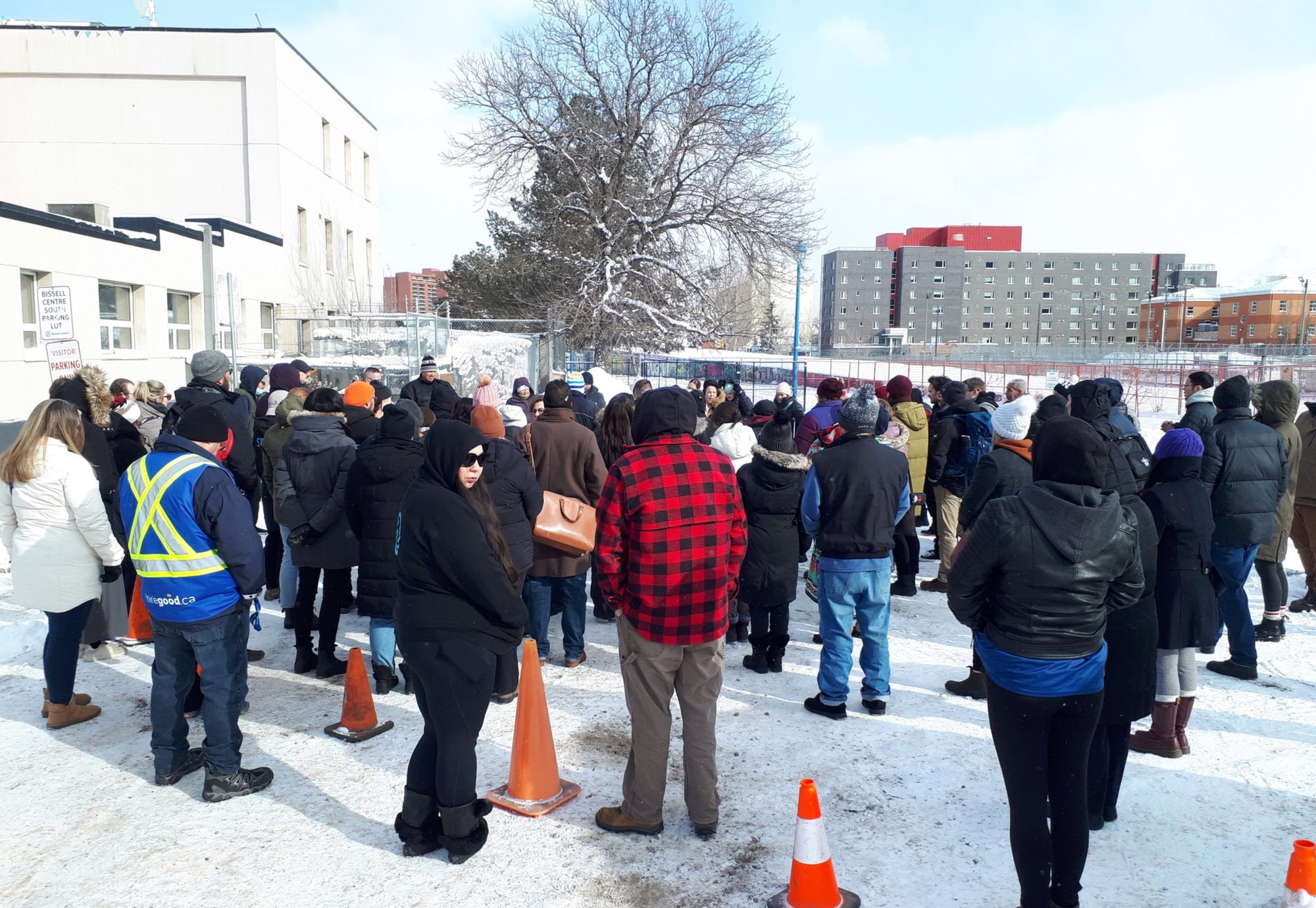 Around 50 people stand outside around a few central speakers giving a memorial, in a parking lot or field.