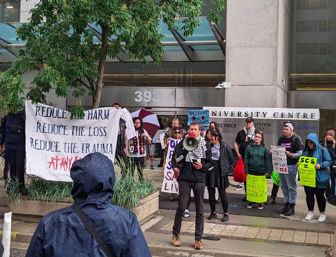 A person speaks into a megaphone beside a sign reading "Reduce the harm. Reduce the loss. Reduce the trauma. At any cost."