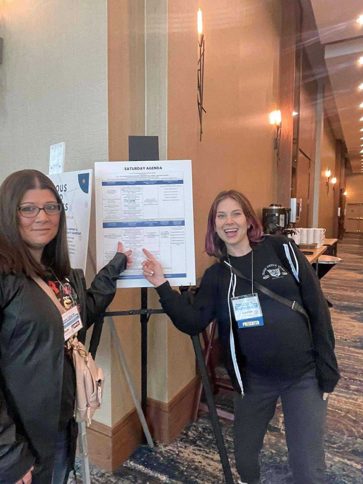 Two women look at the camera with an easel between them holding up a schedule of speaking events. They are pointing at a box that presumably shows their speaking slot. One looks apprehensive and the other has a huge smile. 