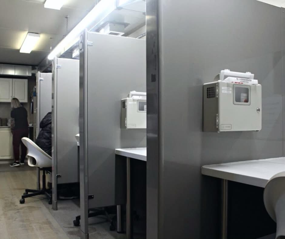 Clean desks with needle discard boxes hung above them, plastic rolling chairs pulled up to them, and one unidentifiable person sitting at one. A woman is in the background preparing something at a countertop. 