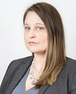 A woman with medium-length light brown hair and blue eyes dressed in professional attire looks neutrally toward the camera. 