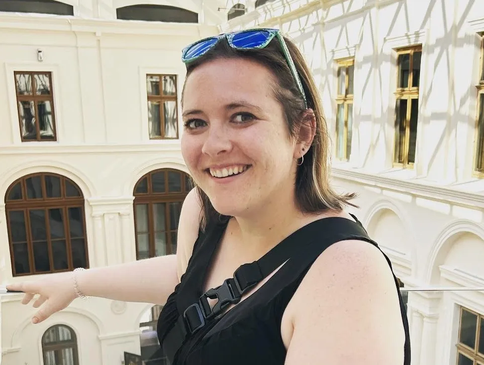 A woman with shoulder-length hair wearing a black tank top and sunglasses on top of her head smiles at the camera from inside an off-white courtyard or atrium. 