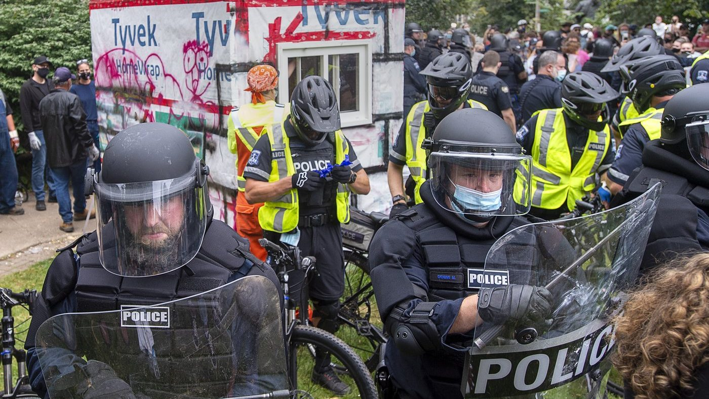 Police in riot gear encircle and defend the removal of a tiny home with people protesting around them. 