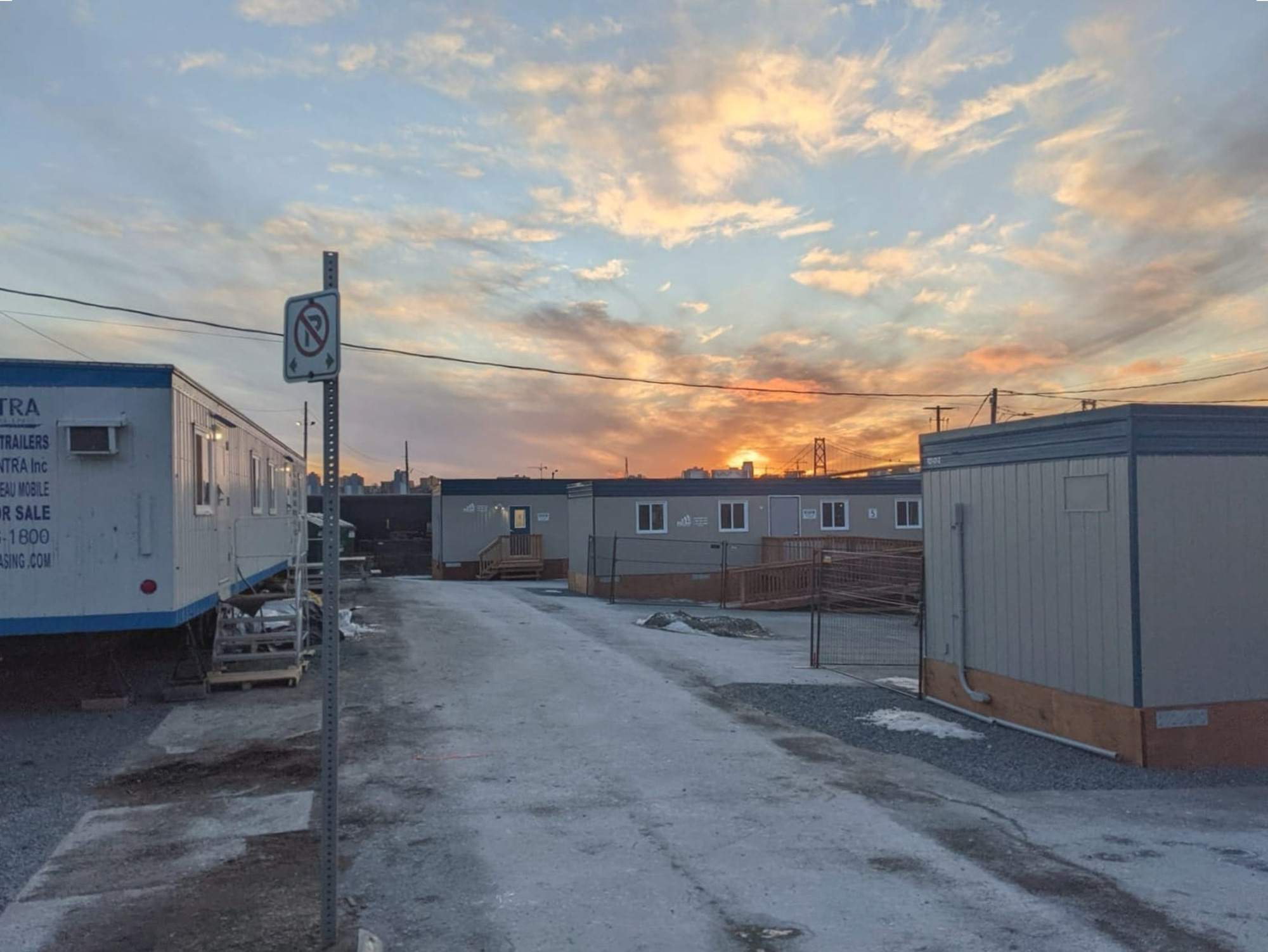 Late-winter scene among several trailers serving as shelter units in a parking lot. A sunset is visible in the background, indicating the direction west. The blue sky is streaked with clouds. 