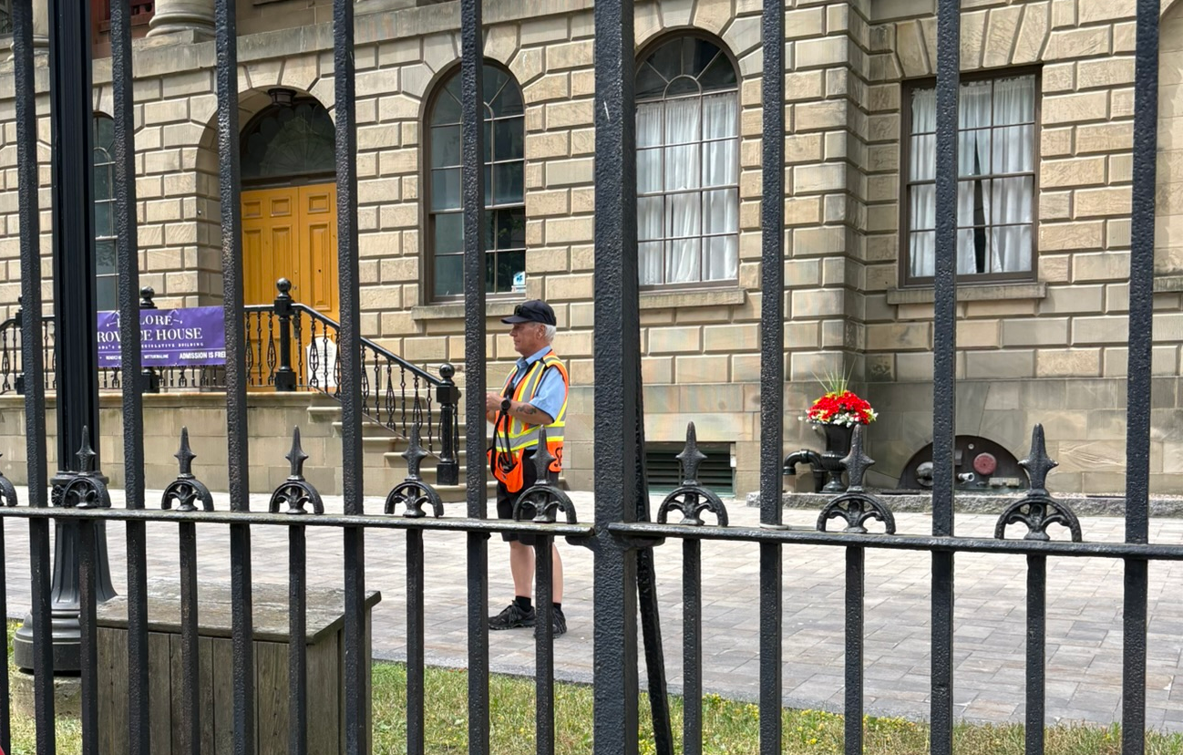 An aging man in a visibility vest takes footage from his cell phone camera, behind a wrought-iron fence.