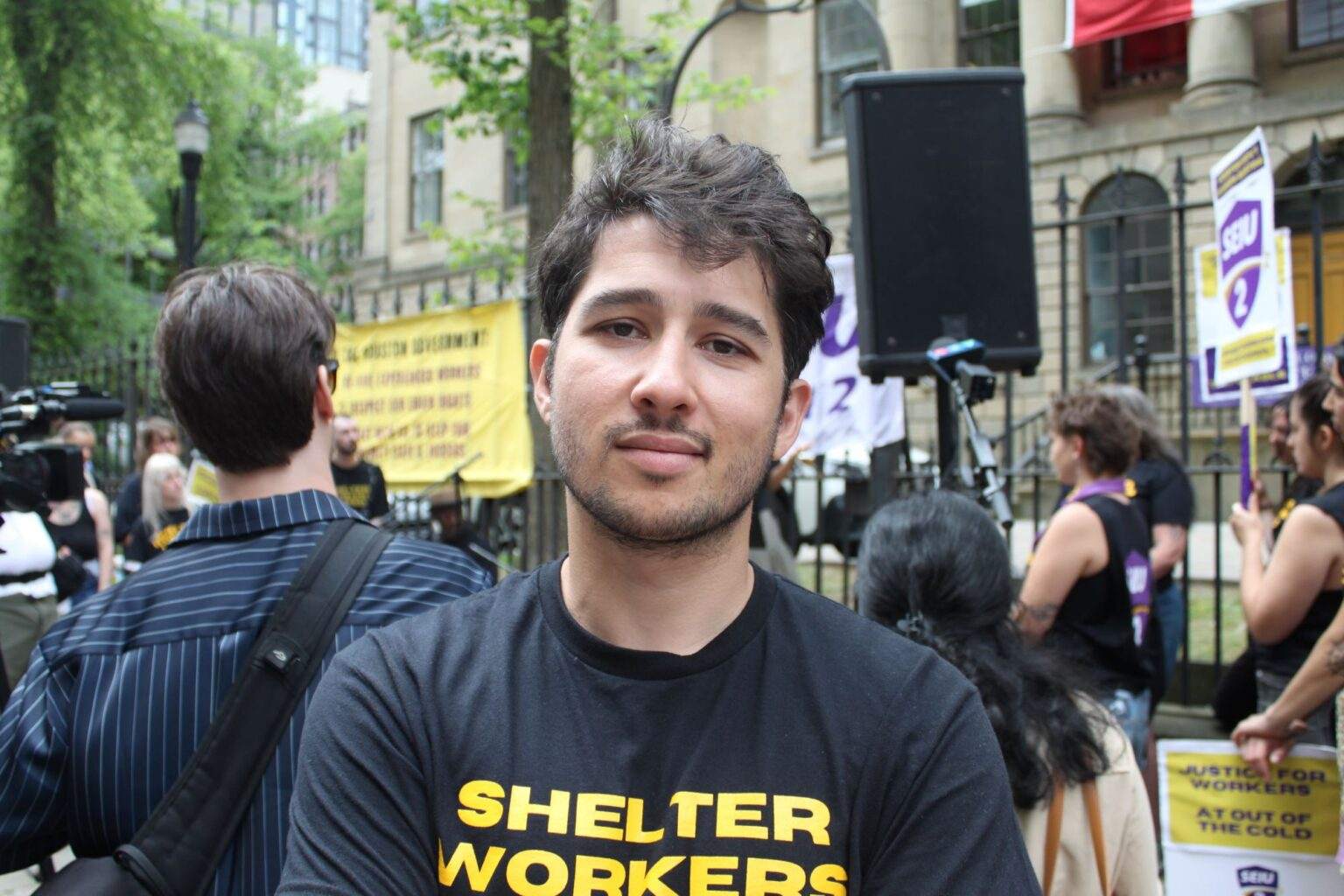 A latinx-presenting man in his twenties wearing a black shirt with yellow lettering that reads "Shelter Workers" faces the camera with a neutral, somewhat saddened expression. Behind him are news cameras and microphones and a crowd of people. 