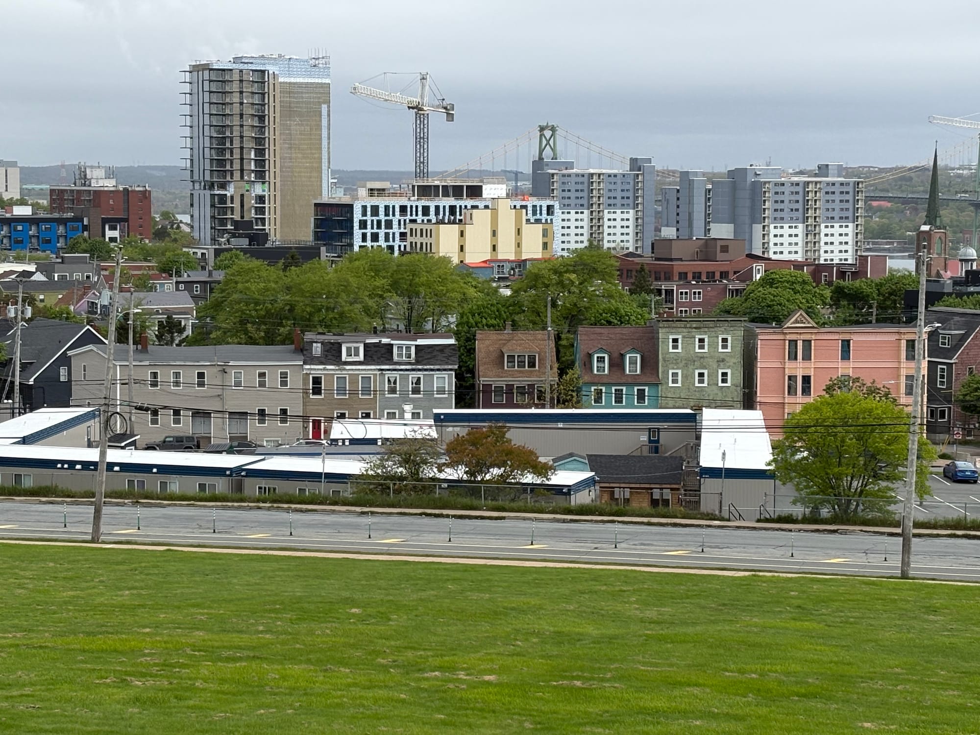 A green space before a road and five modular trailers, then colourful character homes. Beyond that is the cityscape where a construction crane and high rise can be seen.