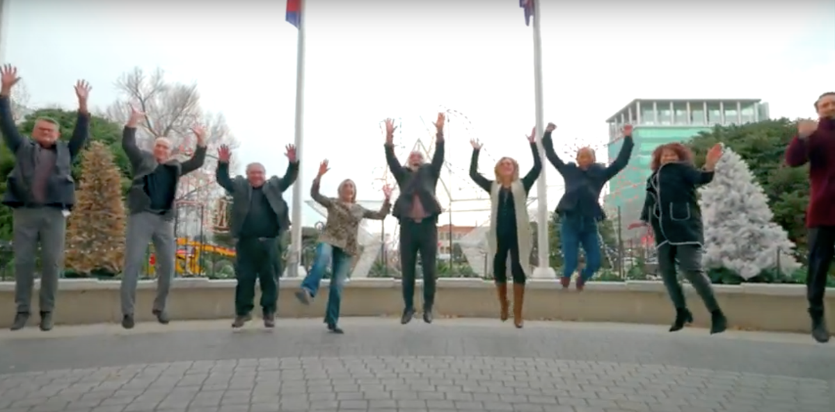 Photo of 9 people in professional attire jumping at the same time in a public space with arms up.  