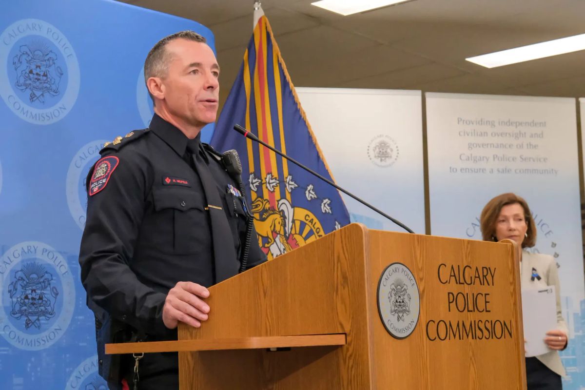 A man with short-cropped hair in a police uniform speaks at a microphone podium.