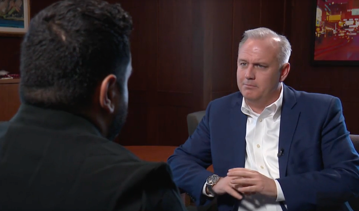 A man in a blue suit jacket and crisp white button shirt folds his hands and gazes cockeyed at an interviewer.