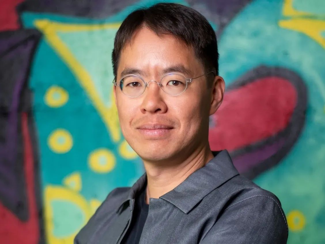 A man with glasses and a collared grey shirt looks stoically at the camera, against a colourful background.