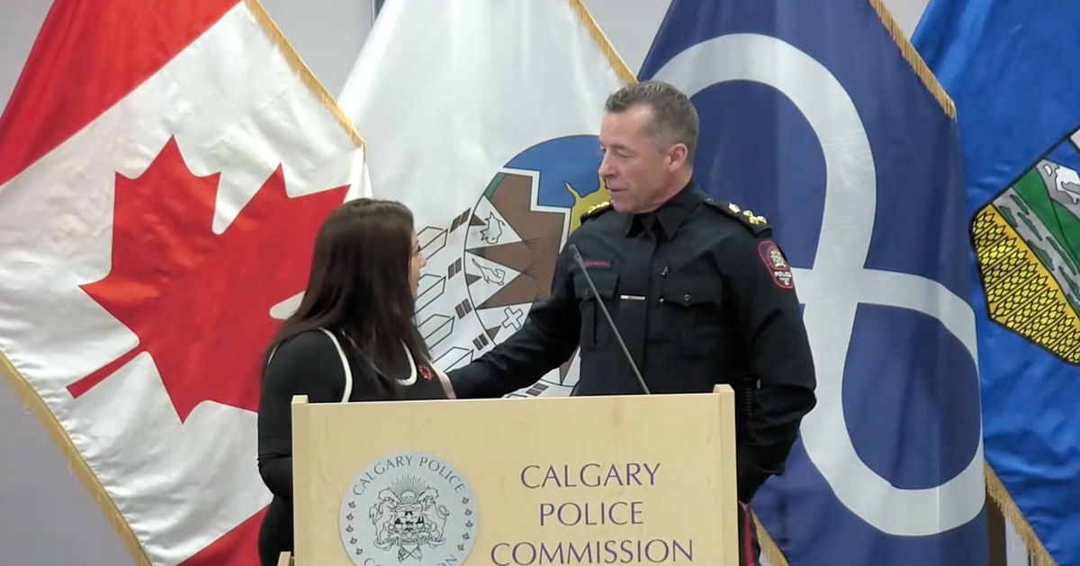 A man in police uniform stands facing a woman with his hand on her upper arm. They are in front of a podium and flags.