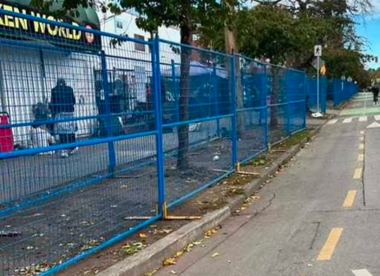 A blue security fence encloses the grassy median between sidewalk and bike lane in Victoria, BC.