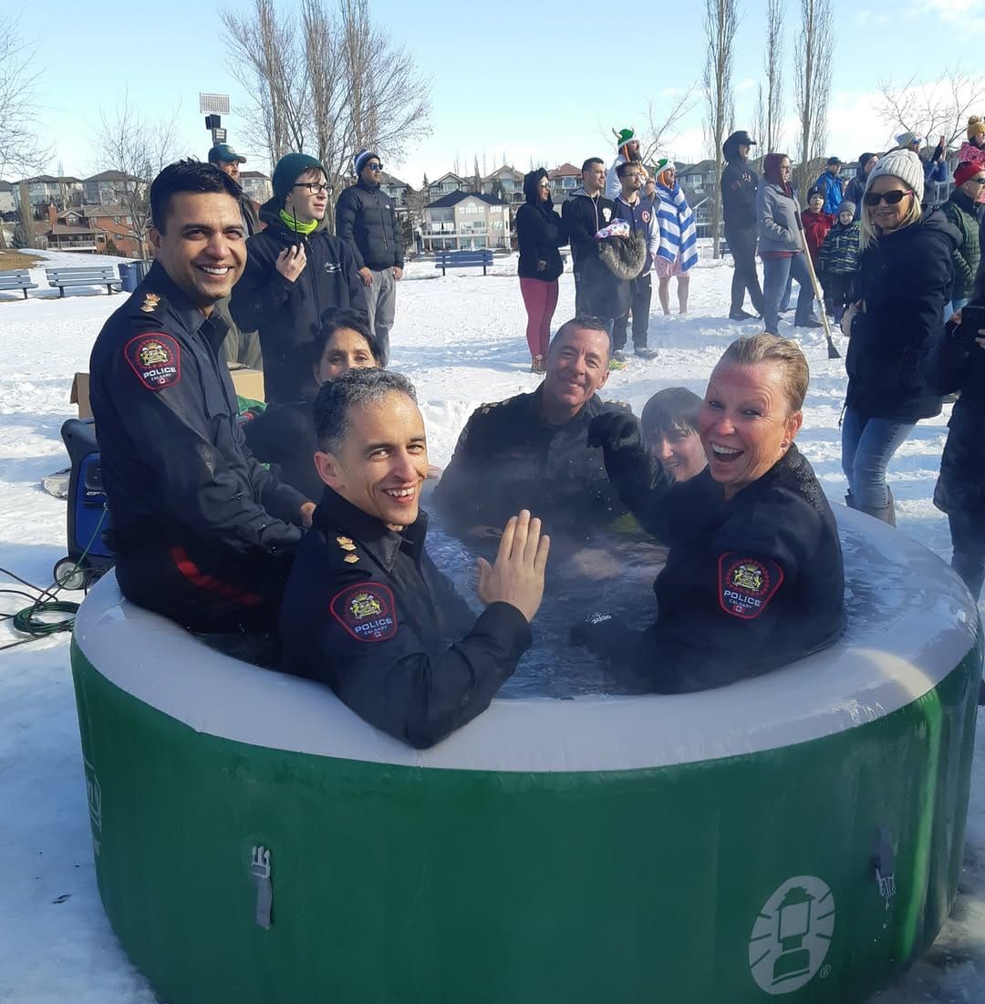 Several police officers in full uniform smile and wave from a small inflatable pool surrounded by snow.