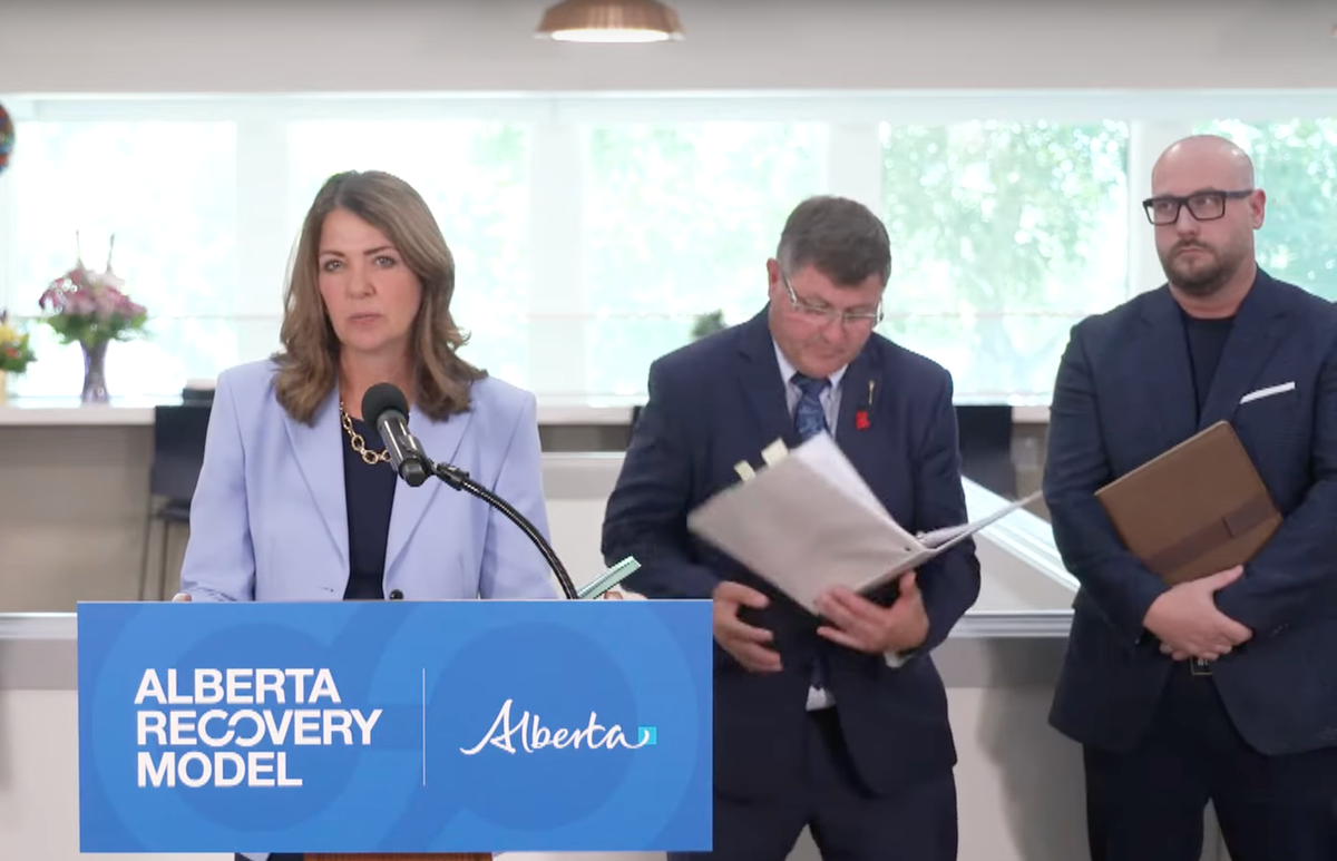 A man in a suit fumbles a binder full of papers behind a woman looking coolly at the camera from a podium.