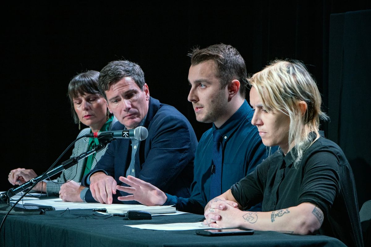 Photo of four people sitting at a black table facing an unseen audience, with microphones in front of their faces.