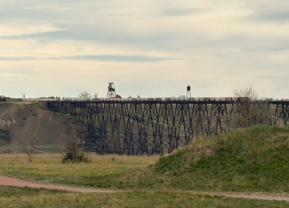 A train exits Lethbridge heading west across the bridge spanning the Oldman River that splits the town in two. The bridge is a few hundred feet high. The sky above is cloudy.