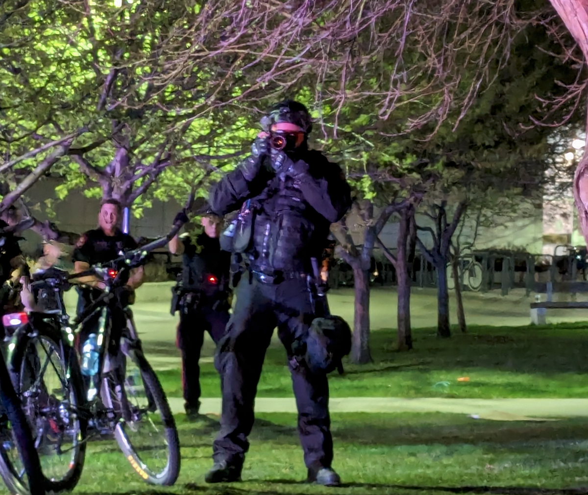 A police officer in a militarized uniform with heltet and body armor holds a camera to his face, to photograph protesters at a night-time protest in a field.