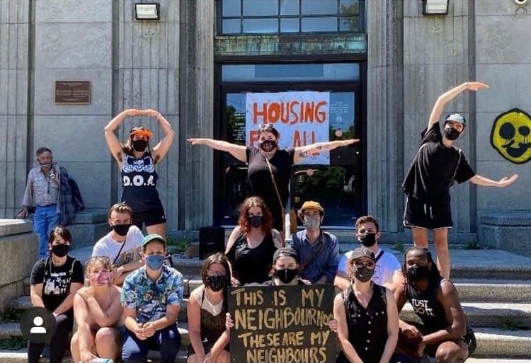 About fifteen people gather in front of a polished marble building, three of them forming the letters OTC. Others hold protest signs about housing for all. 