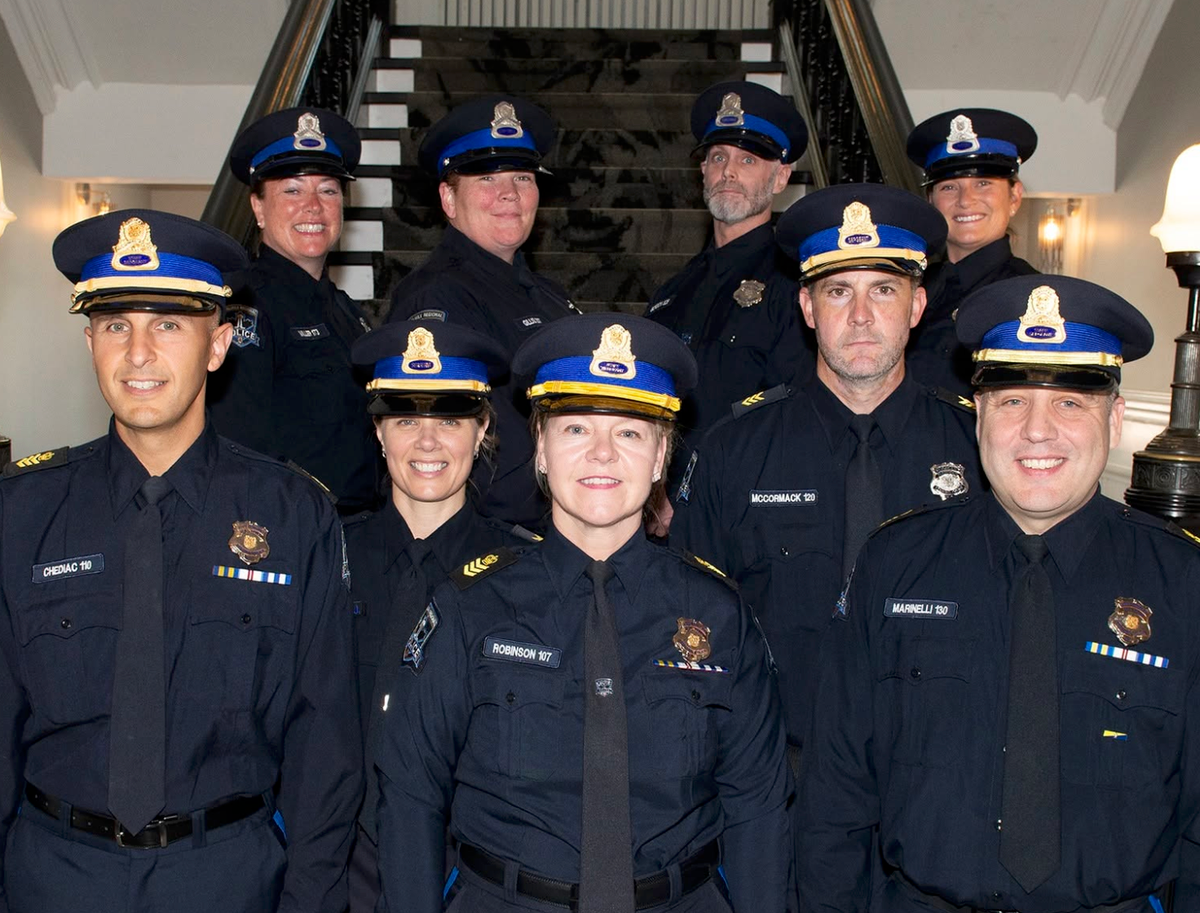 Nine police officers in police caps and uniforms smiling in front of an old staircase.