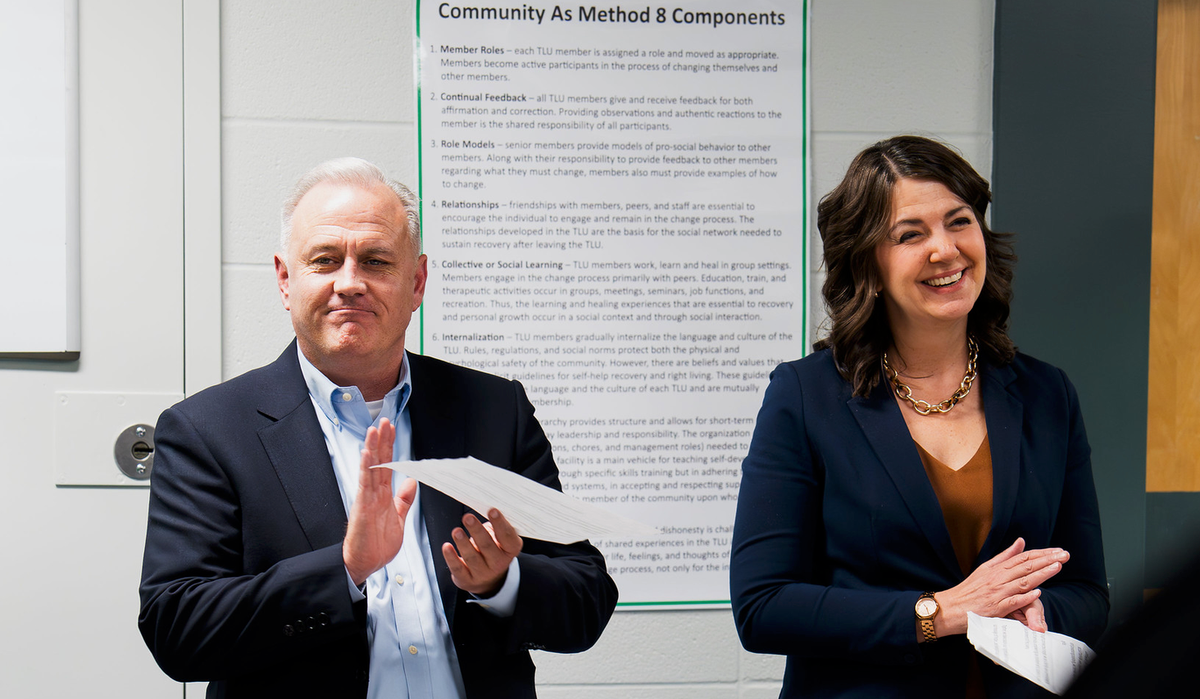 A man and a woman, both in suits, clap and smile in a cinder block room inside a jail. 