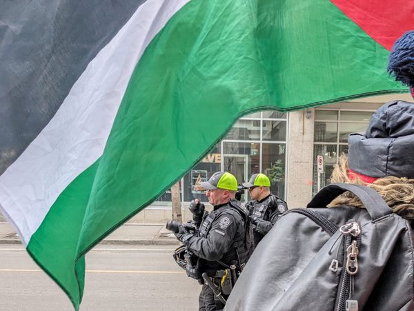 A Palestinian flag hangs over the heads of two police officers monitoring protesters with cameras.