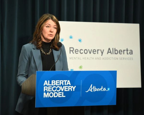 A woman is in mid-sentence behind a podium that reads "Alberta Recovery Model" and in front of a screen reading "Recovery Alberta."
