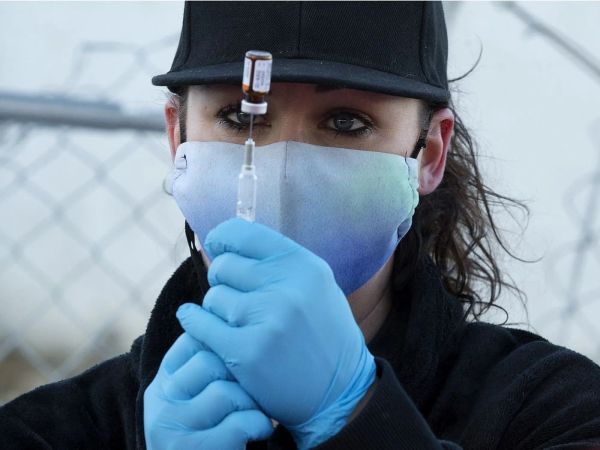A woman wearing mask and gloves stares intently at an inverted vial and syringe she is holding in front of her face.