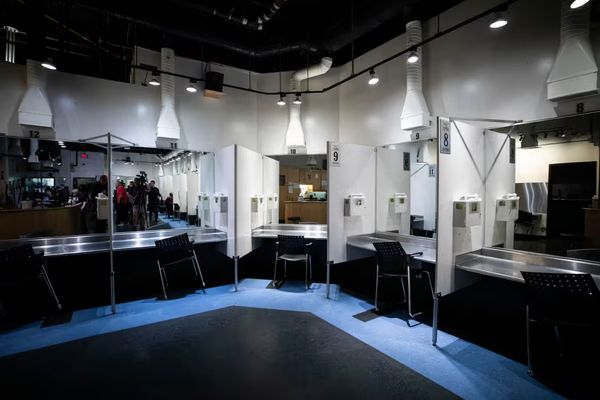 A darkened room with an L-shaped row of clinical-looking seated booths in front of mirrors. Above each booth is a vent, and beside each one is a sharps disposal bin. 