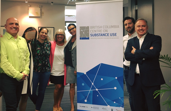 Seven people posing and smiling in front of an advertisement poster reading "BC Centre on Substance Use".