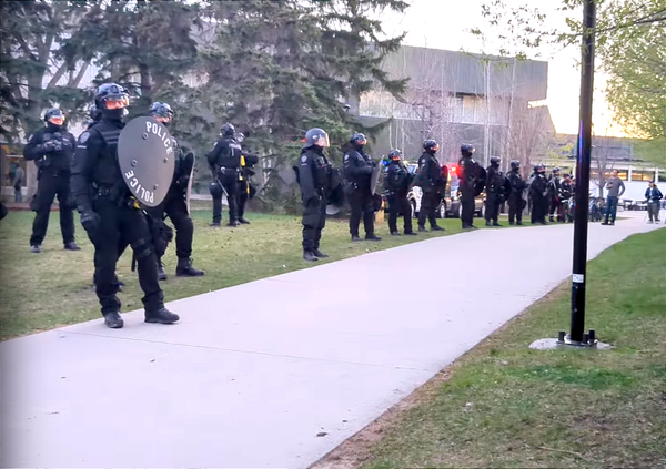 A line of police in riot gear with shields, helmets, batons and pepper bullet guns at a university campus.
