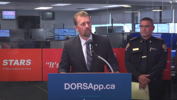 A man in a suit looks earnestly in the direction of the camera while standing at a podium with "DORSapp.ca" written on it.