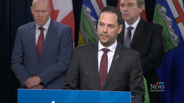 A man in a suit looks toward an unseen crowd from a podium in front of provincial and national flags. Two men also in suits look on from behind.